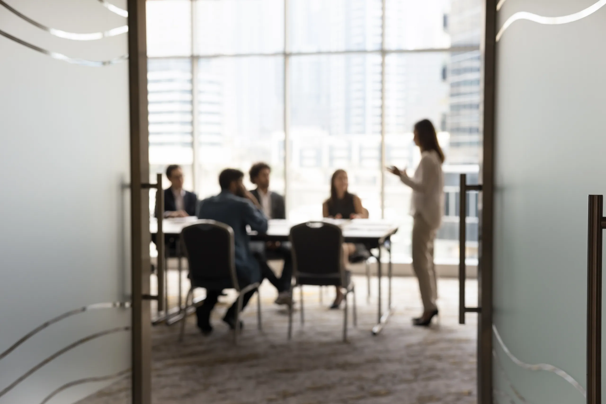 A leadership team meeting around a boardroom table, seen through open frosted-glass doors